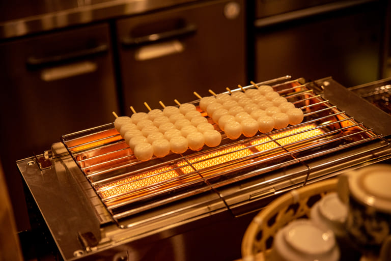 焼き串だんごが並ぶグリル台と暖色の照明に照らされたキッチン風景