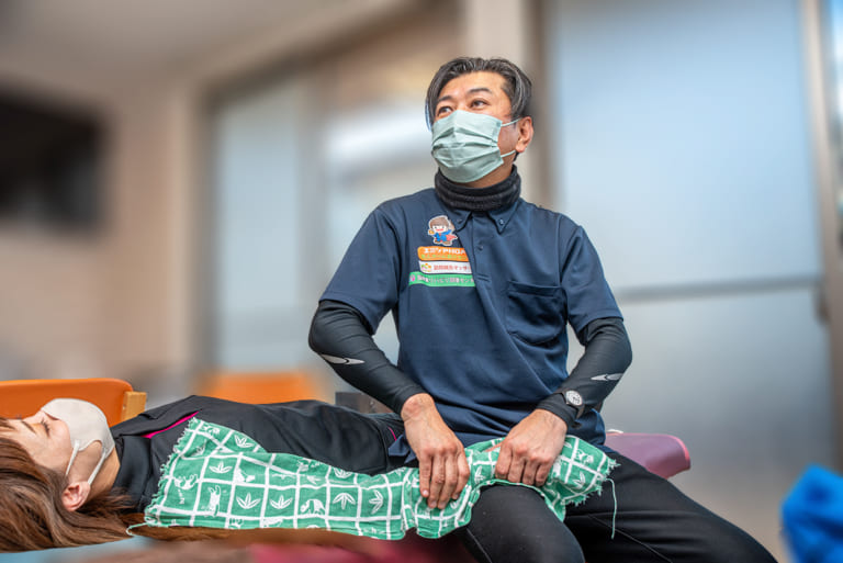 訪問マッサージを実演する男性セラピストが、患者を横向きにして腰回りをマッサージしている場面。