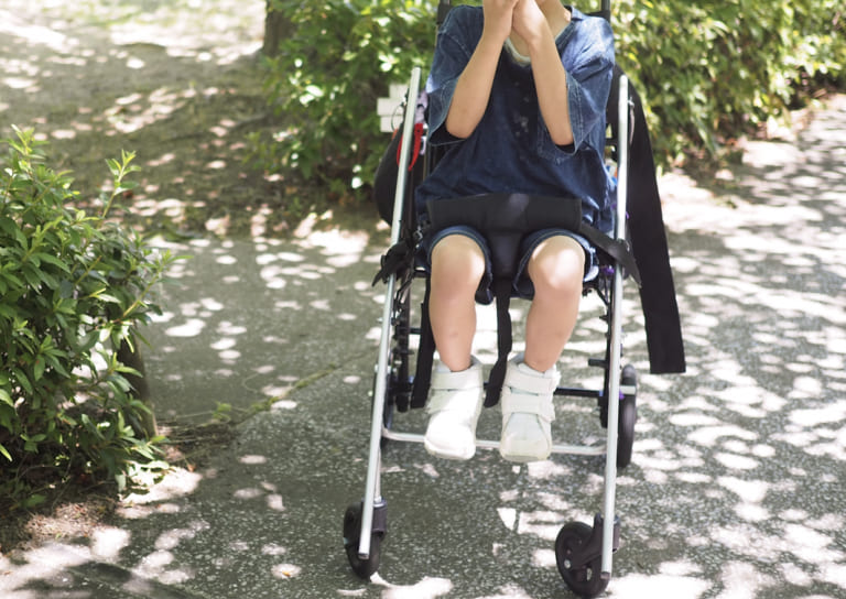 公園の道を車椅子で移動する子どもが手で顔を覆っている様子