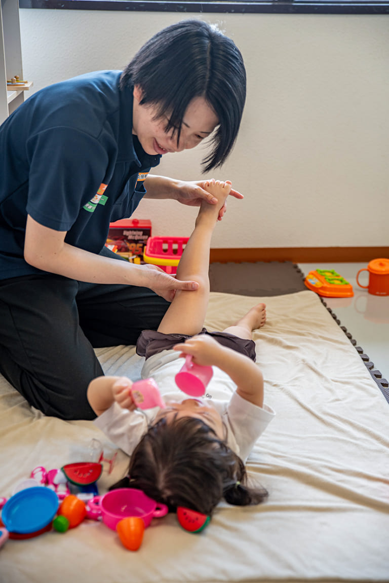 居室で訪問マッサージを行い、子どもの足を優しく動かしてリハビリをサポートする施術者