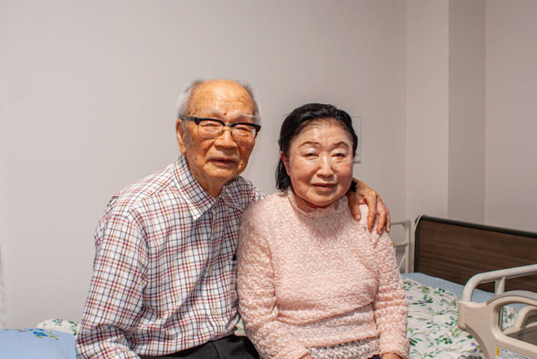 訪問リハビリマッサージを受ける高齢夫婦が病室のベッド横で寄り添う様子