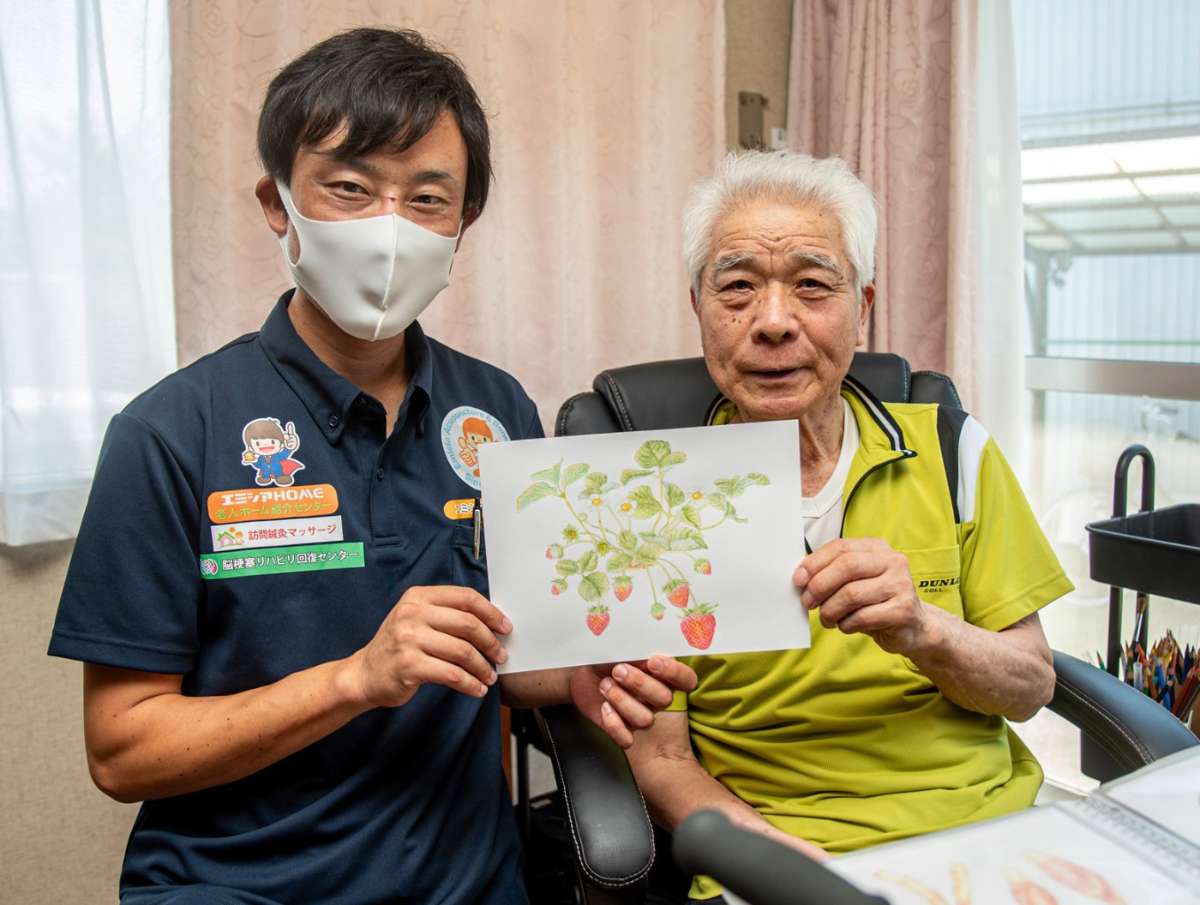 手作りのイチゴの絵を見せる高齢男性と訪問マッサージ担当者の記念ショット